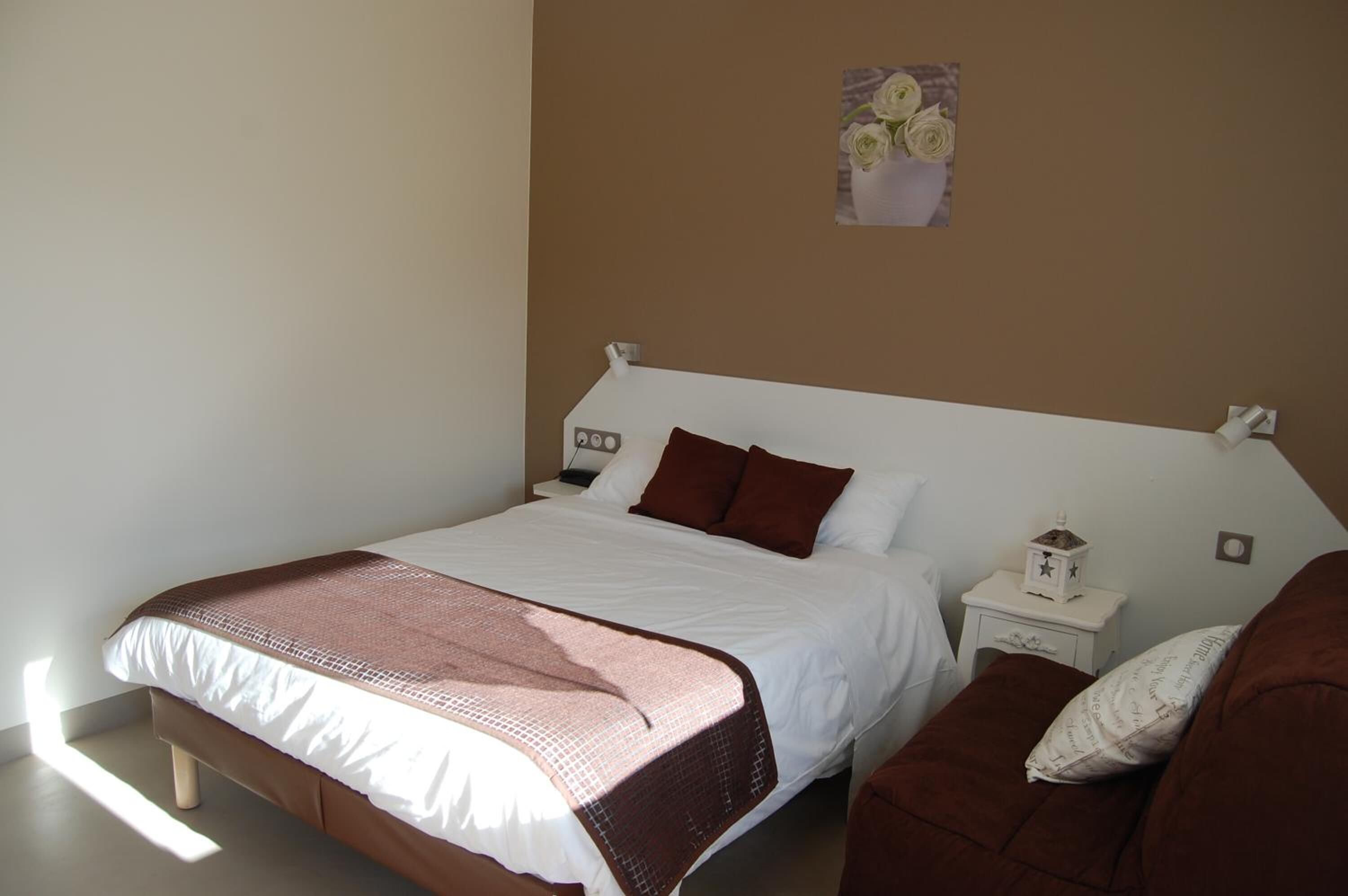 A neatly made bed with a white and brown bedspread, a small bedside table with a lamp, and a wall-mounted picture of flowers.