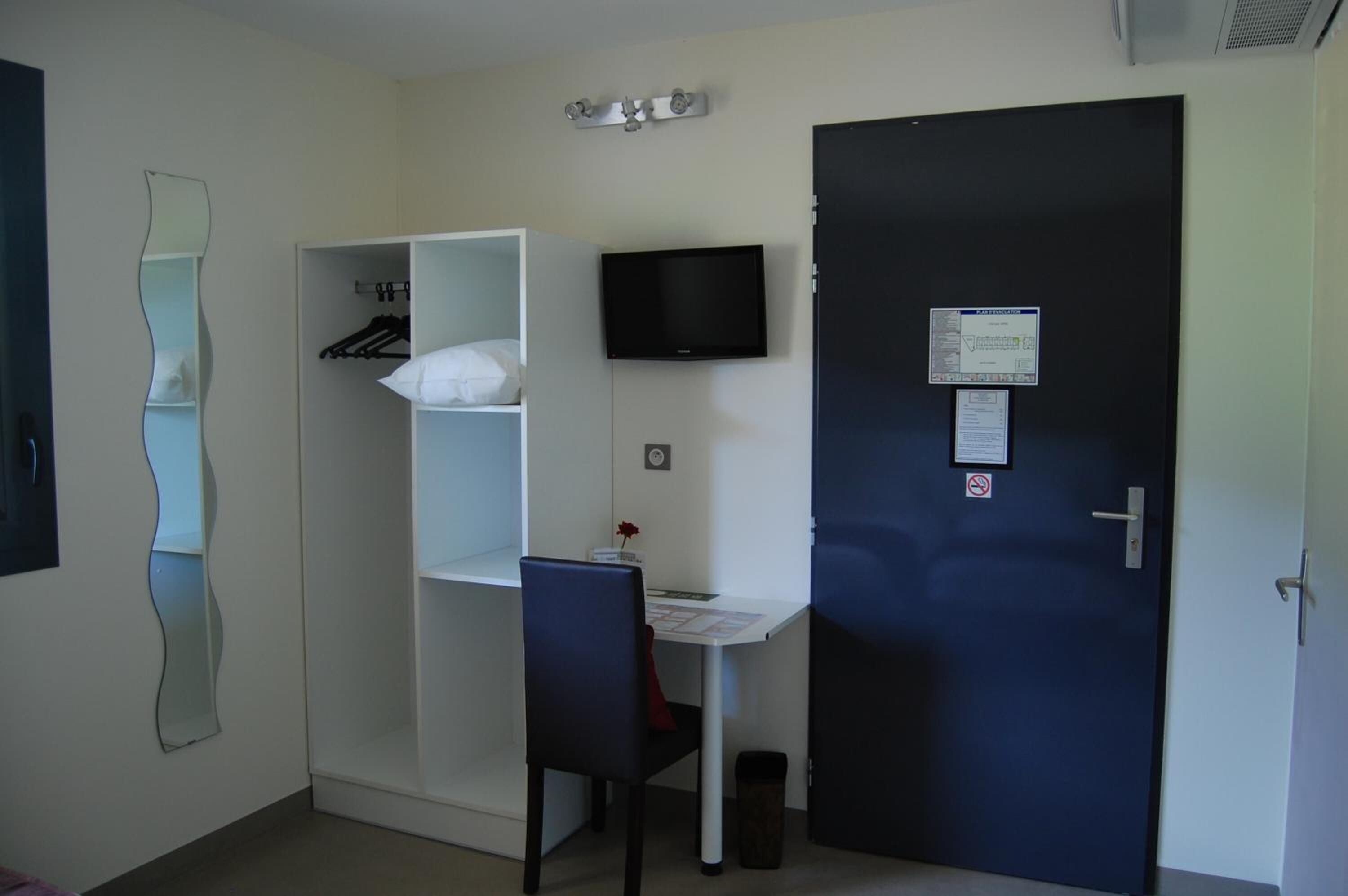 Una habitación pequeña con armario, escritorio, silla, televisor y una puerta con un aviso.