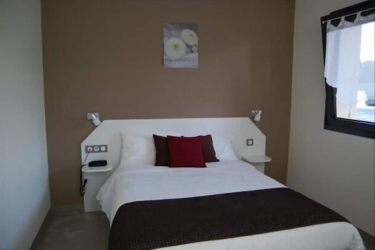 A hotel room with a bed, a nightstand, a wall-mounted power outlet, and a window with a curtain.
