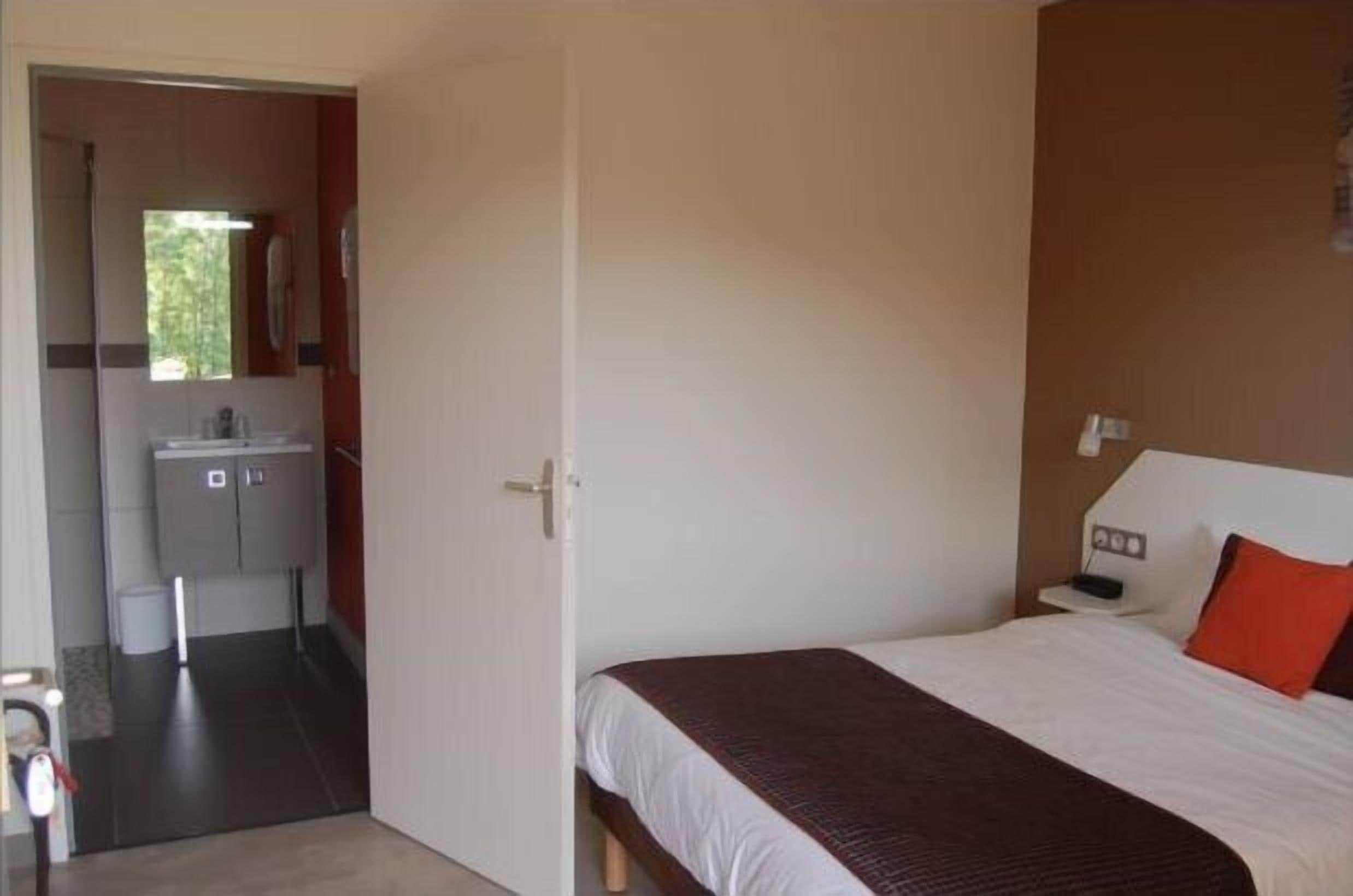 A hotel room with a bed, a nightstand, a red pillow, and a bathroom visible through an open door.