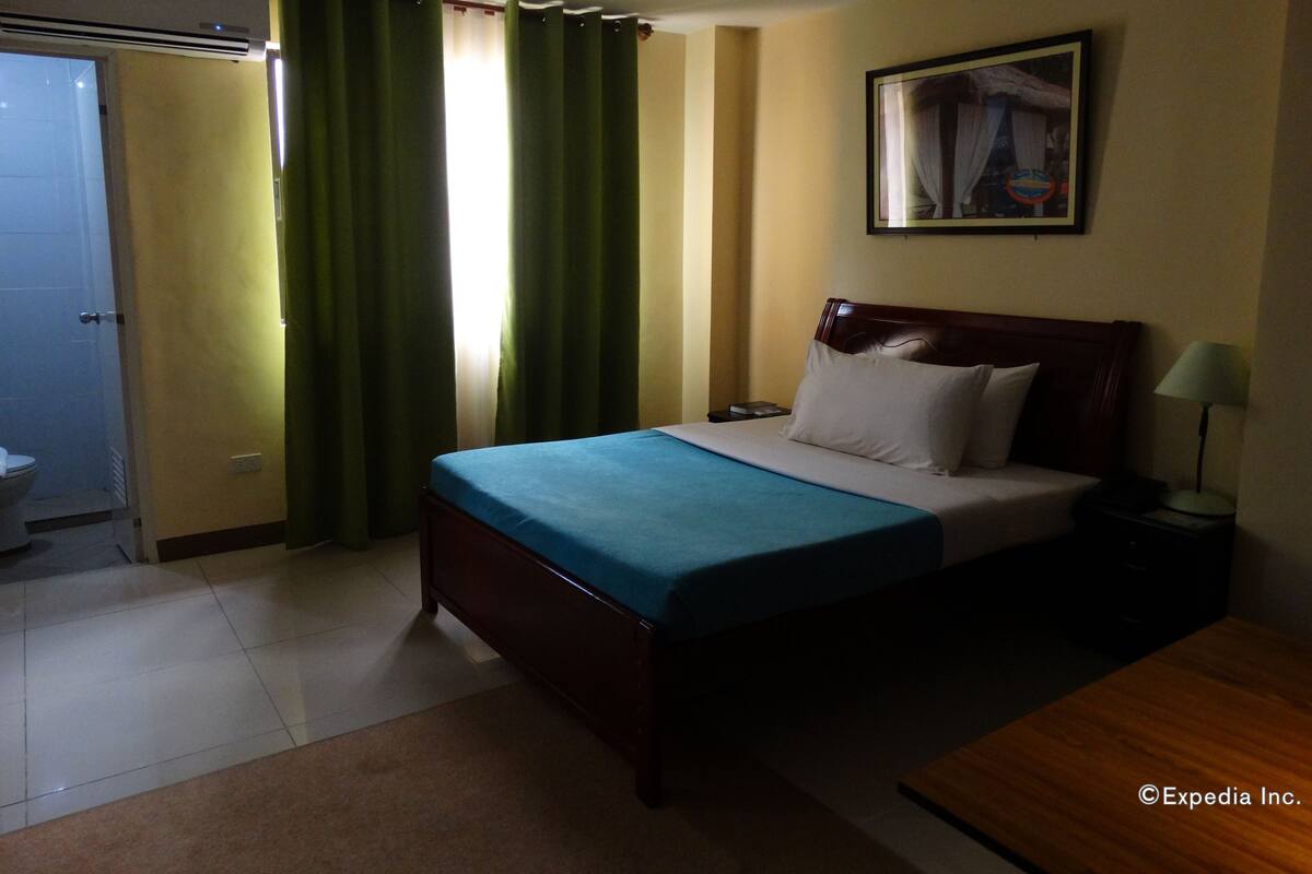 A hotel room with a bed, a nightstand, a lamp, a painting on the wall, and a bathroom with a toilet and shower.