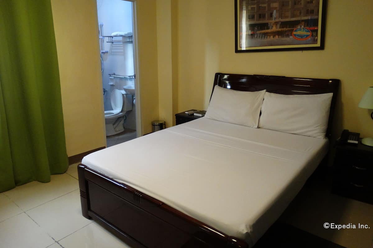 A hotel room with a bed, a nightstand, a lamp, a framed picture on the wall, and a bathroom visible through an open door.