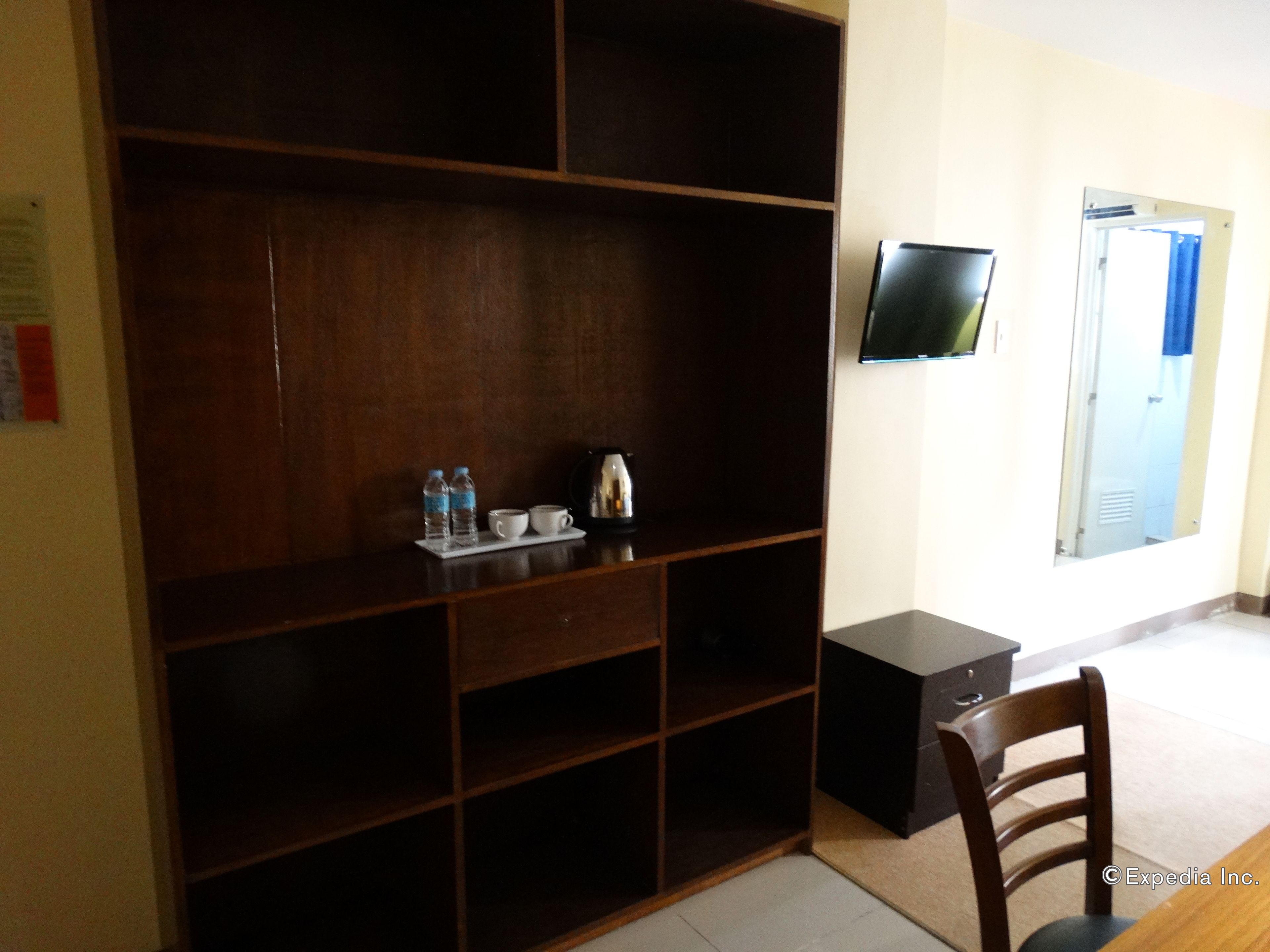 A hotel room with a large wooden wardrobe, a small table with a kettle and cups, a television on the wall, and a chair.