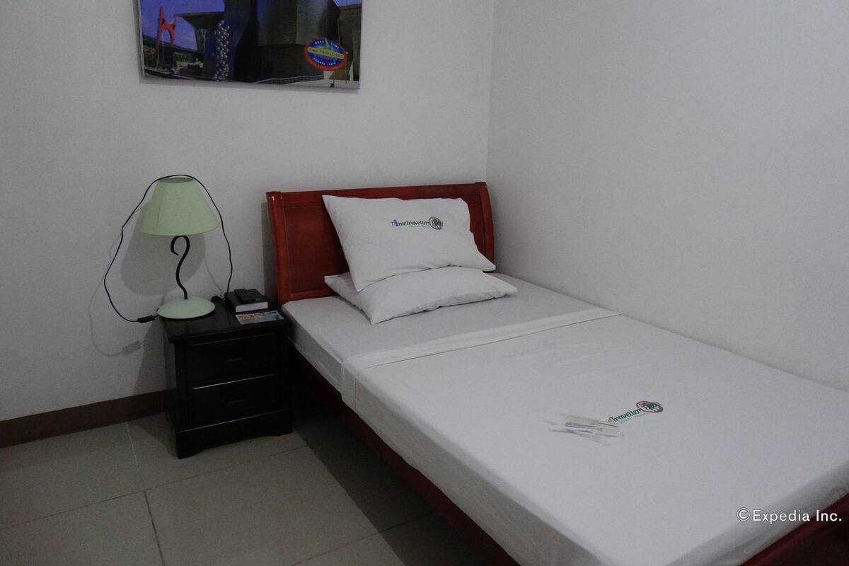 A hotel room with a single bed, a nightstand with a lamp, a painting on the wall, and a small table with a remote control.