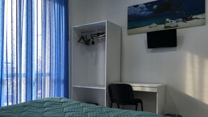 A bedroom with a bed, a desk, and a chair. There is a wardrobe with clothes hanging, a painting on the wall, and a window with curtains. - Top Floor (Naples)