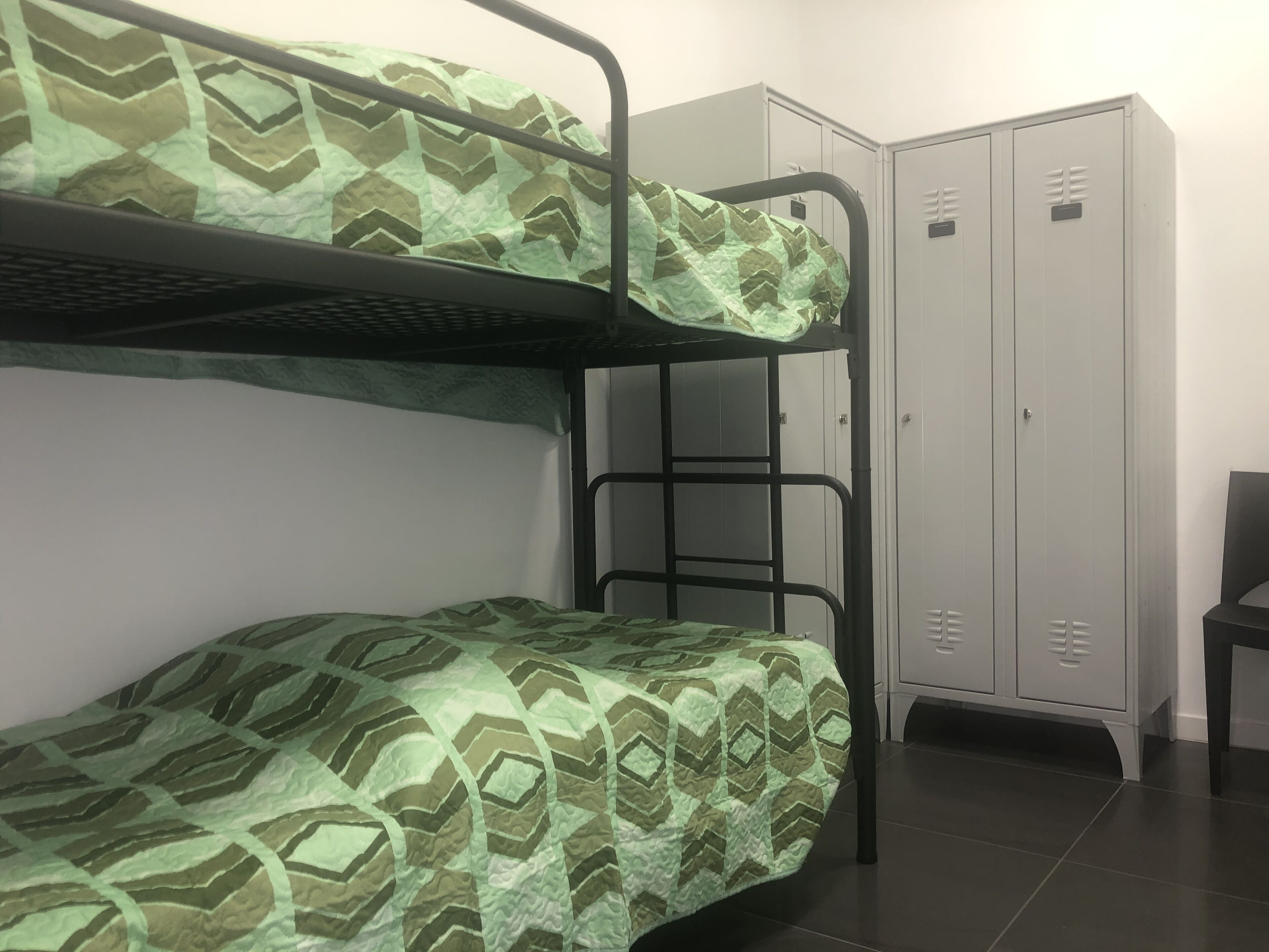 A bunk bed with a patterned green and brown cover, a metal bunk bed frame, and a set of grey lockers against a white wall.