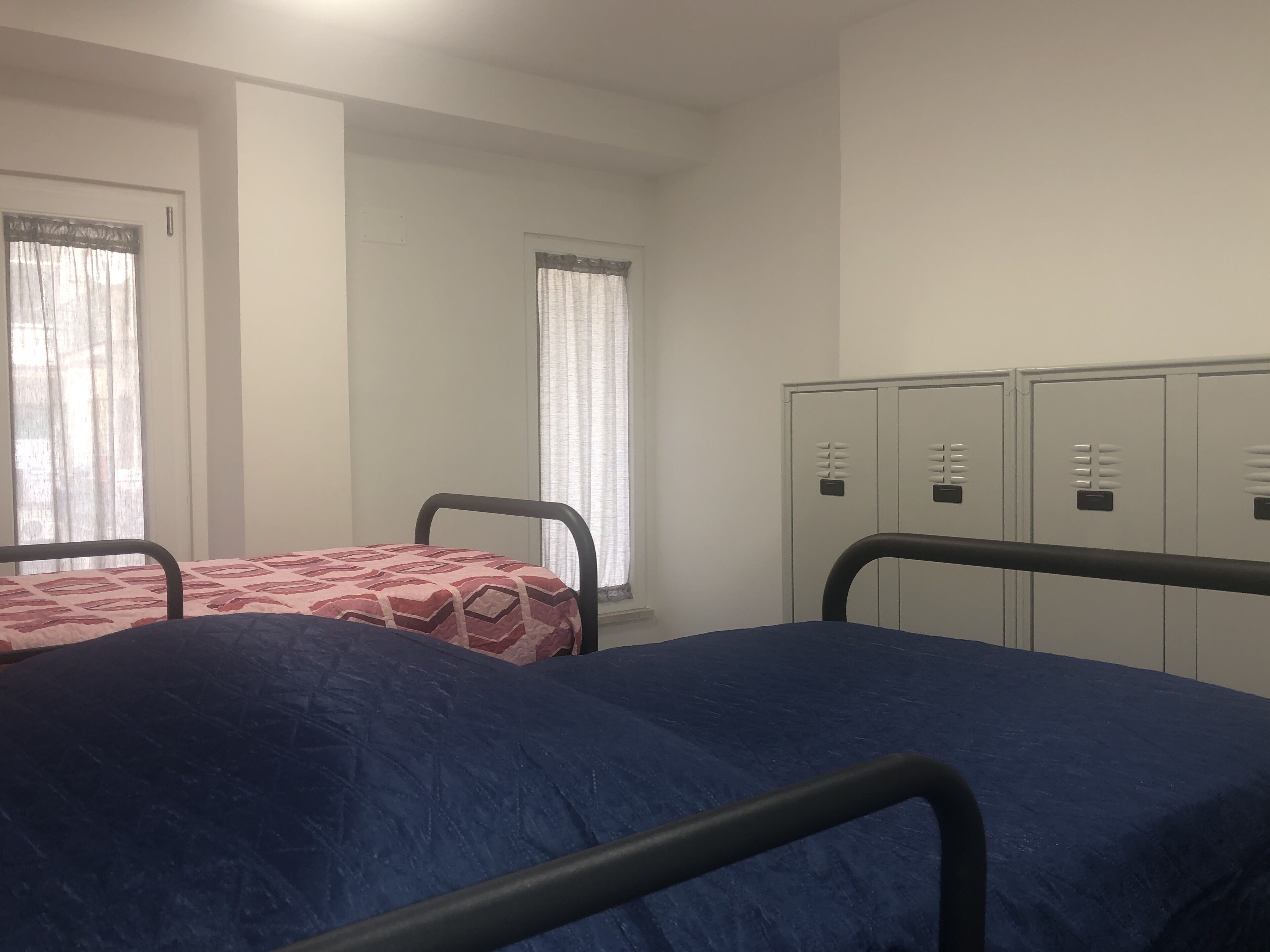 A dormitory room with two beds, a window with curtains, and a row of lockers.