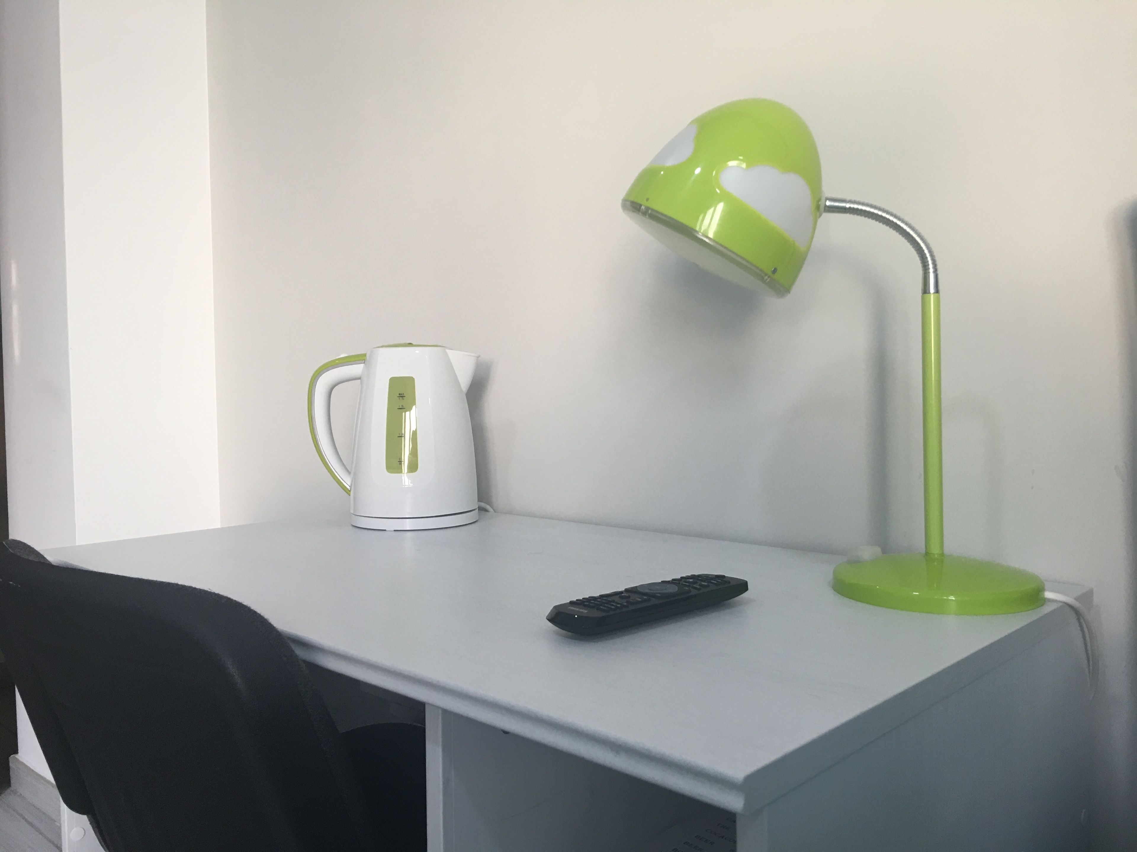 A white desk with a green and white kettle, a green desk lamp, and a black remote control.