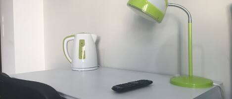 A white desk with a green and white kettle, a green desk lamp, and a black remote control.