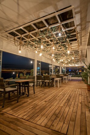 An outdoor dining area with wooden flooring, tables, and chairs, featuring a hanging light fixture and large windows. - Side Beach Club Hotel (Manavgat)
