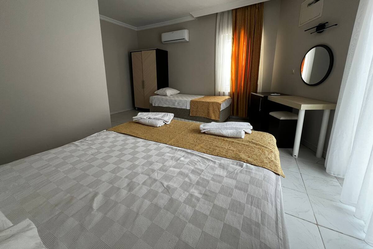 Habitación de hotel con una cama grande, dos camas más pequeñas, un escritorio, un armario, un equipo de aire acondicionado y un espejo.