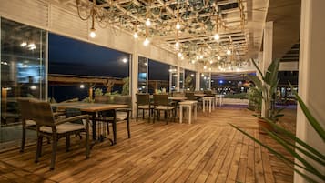 An outdoor dining area with wooden flooring, wicker chairs, and tables. There are large windows along the walls, and a ceiling with hanging lights.