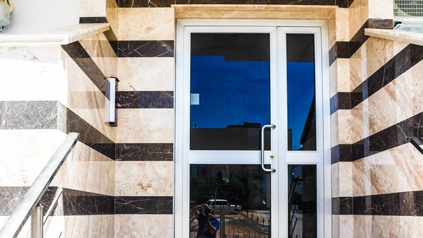 Entrada de um edifício com porta de vidro, corrimão de metal e piso estampado.