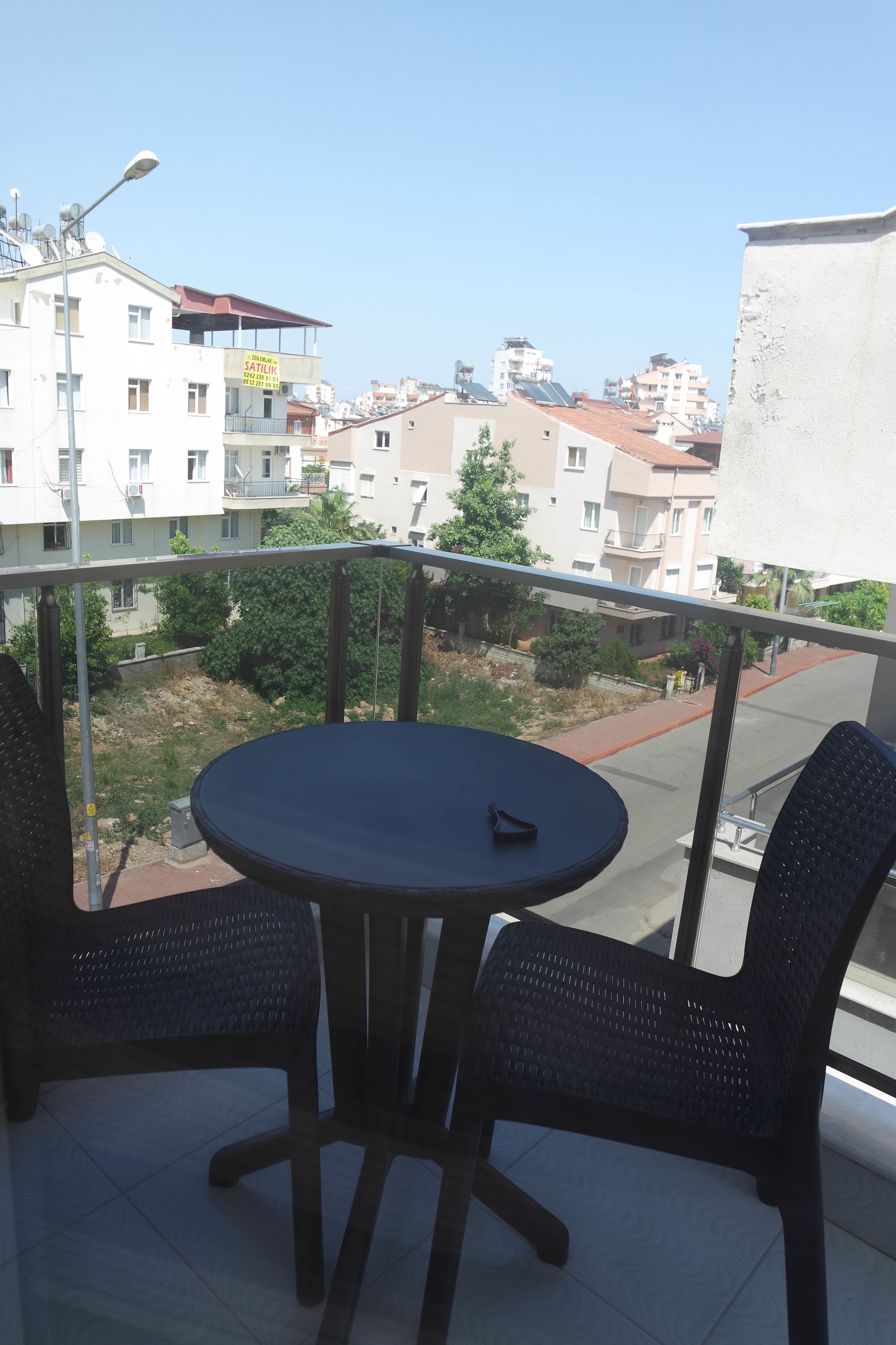 A balcony with a round table and two chairs, overlooking a residential area with buildings and a clear sky.