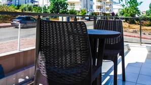 Varanda com duas cadeiras de vime e uma mesa, com vista para uma rua com edifícios e um carro estacionado.