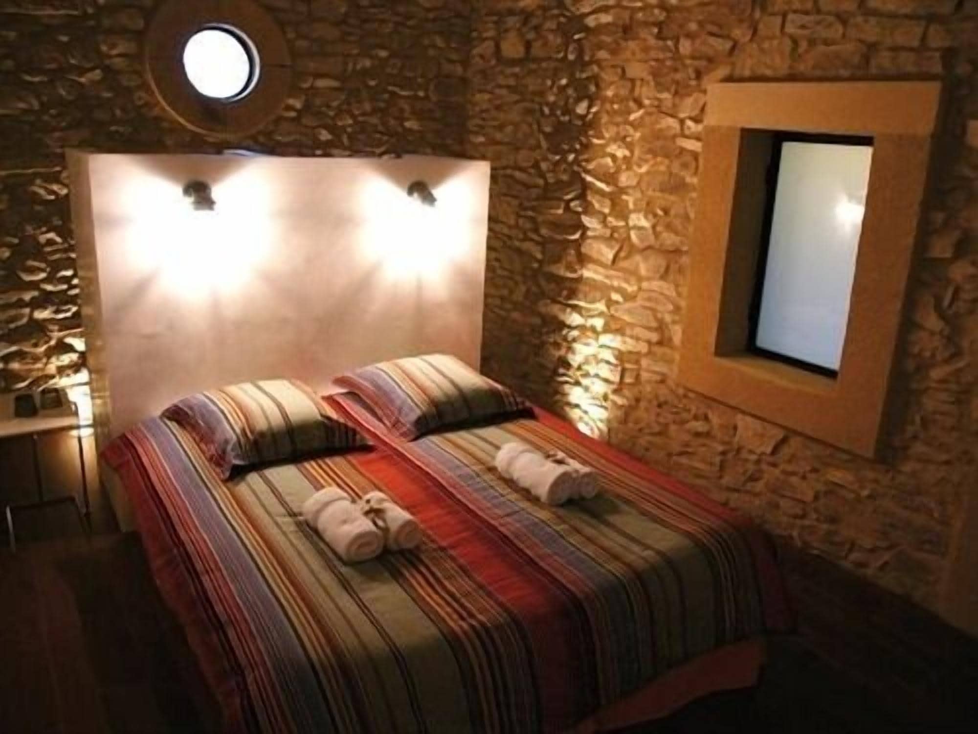 Een kamer met een stenen muur, een bed met gestreepte lakens, twee opgerolde handdoeken, een nachtkastje en een ronde spiegel.