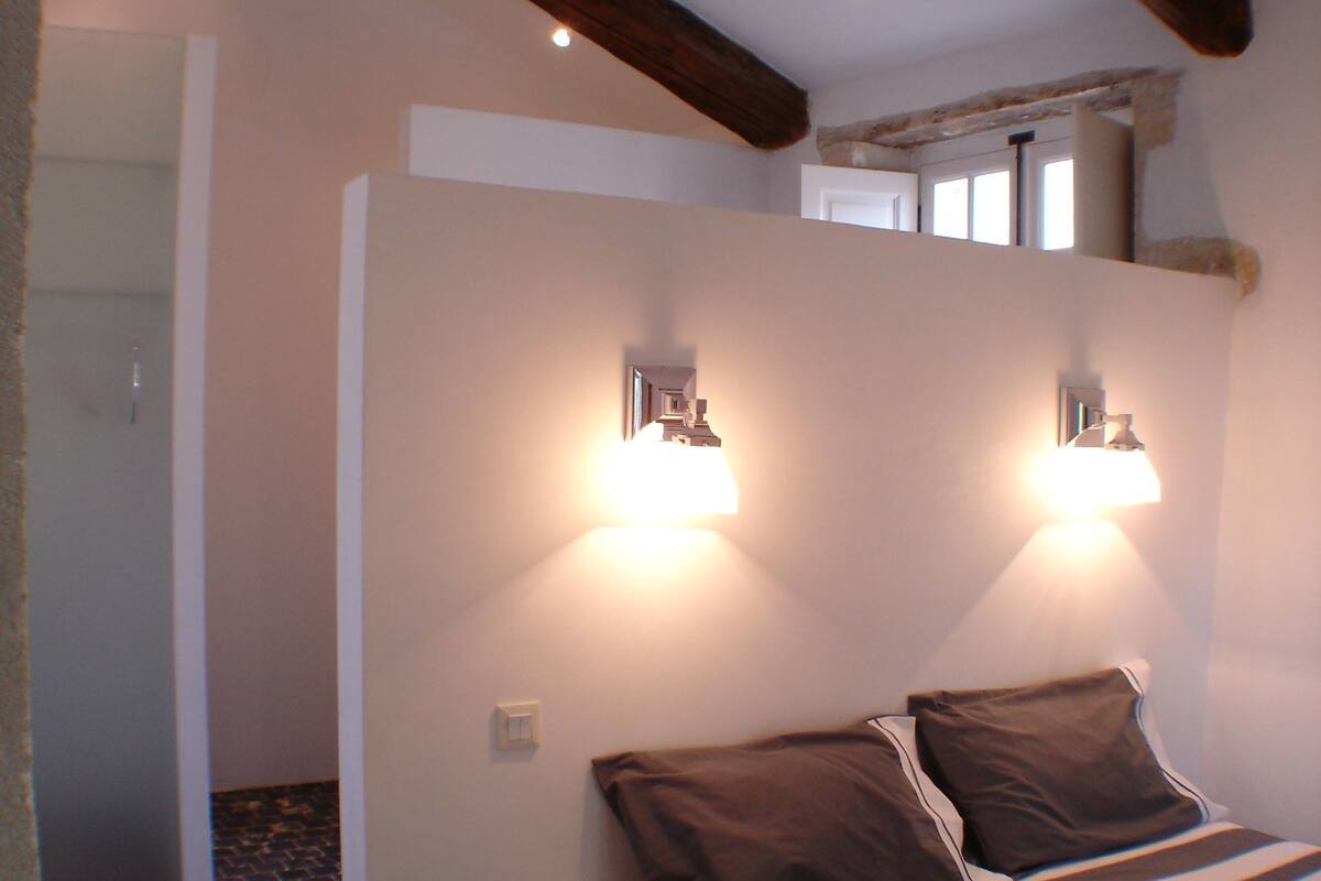 A bedroom with a bed, two wall-mounted lights, a brick-patterned floor, and exposed wooden beams.