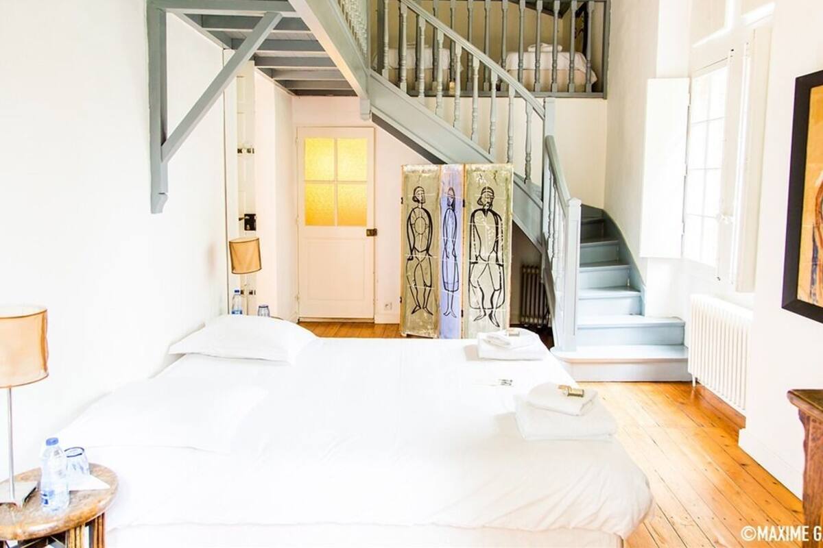 A hotel room with a bed, a nightstand, a lamp, a water bottle, a towel, a staircase, and a painting on the wall.