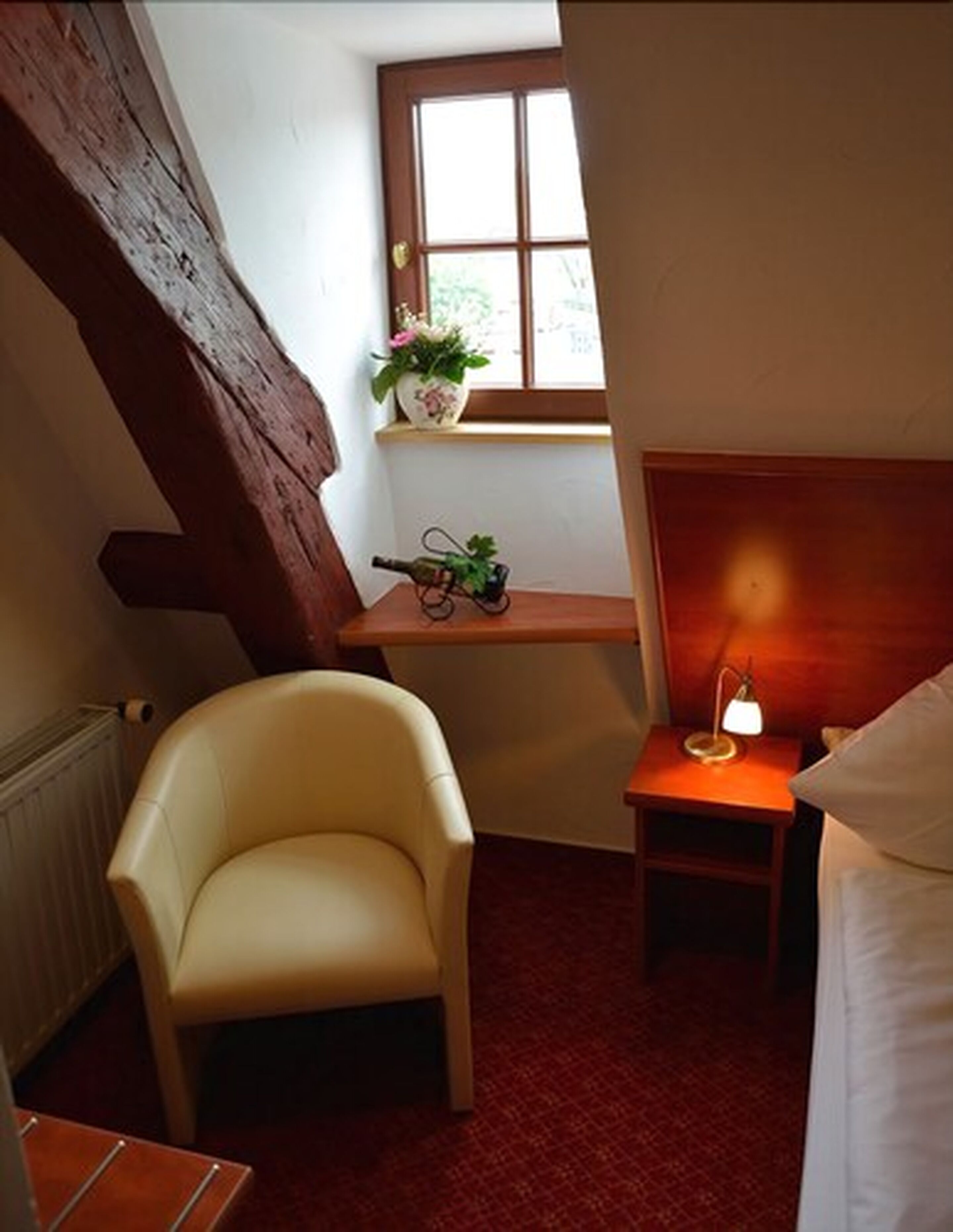 Ein kleiner, gemütlicher Raum mit schrägen Wänden, einem Sessel, einem Nachttisch mit Lampe und einem Fenster mit Blick ins Grüne.