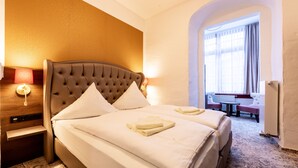 Une chambre d’hôtel avec un grand lit, une table de chevet avec une lampe et une vue sur un espace de restauration, visible à travers une porte en arc de cercle.