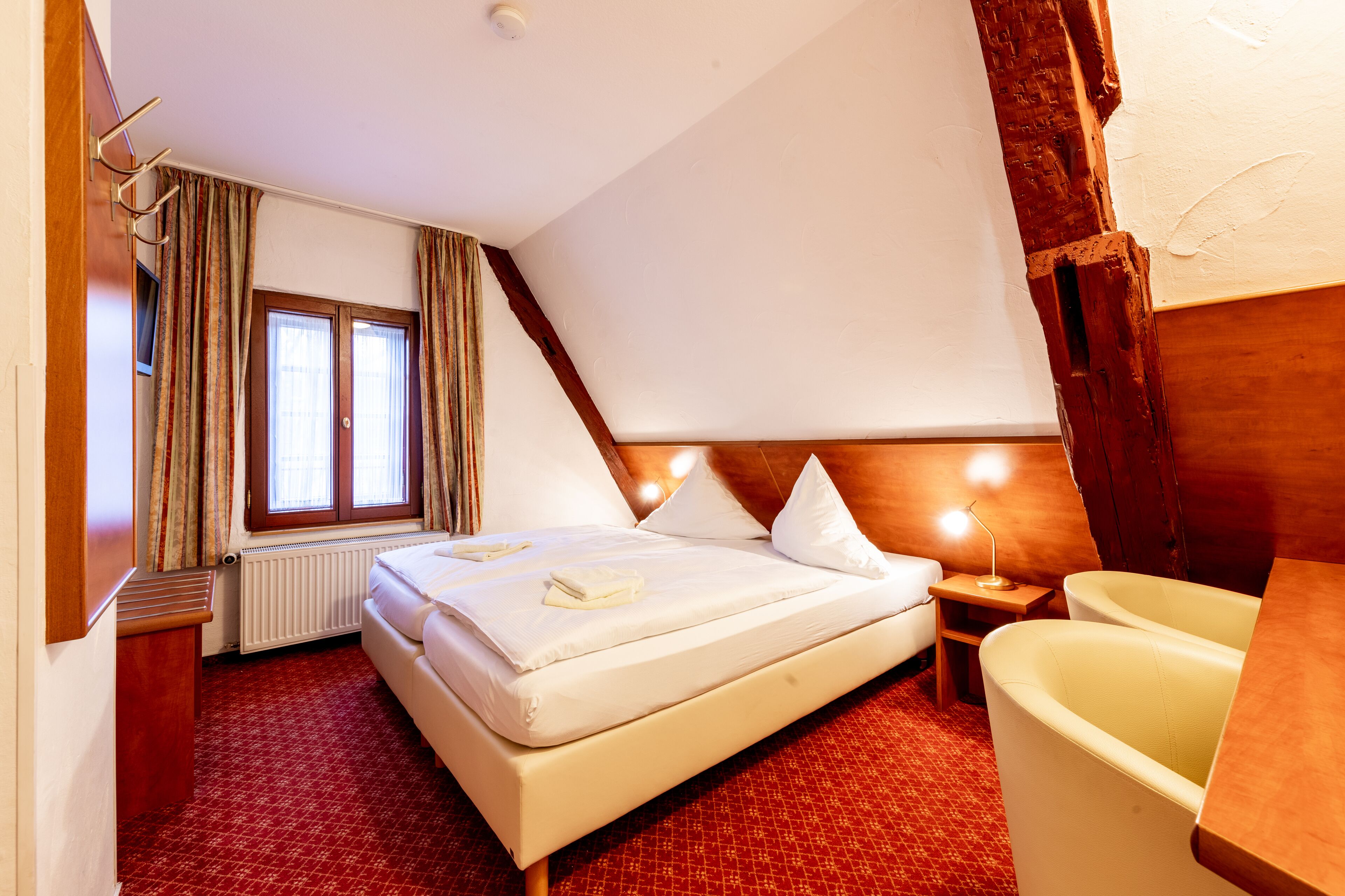 Habitación de hotel con cama, ventana con cortinas, radiador, cabecera de madera, mesita de noche con lámpara y bañera.
