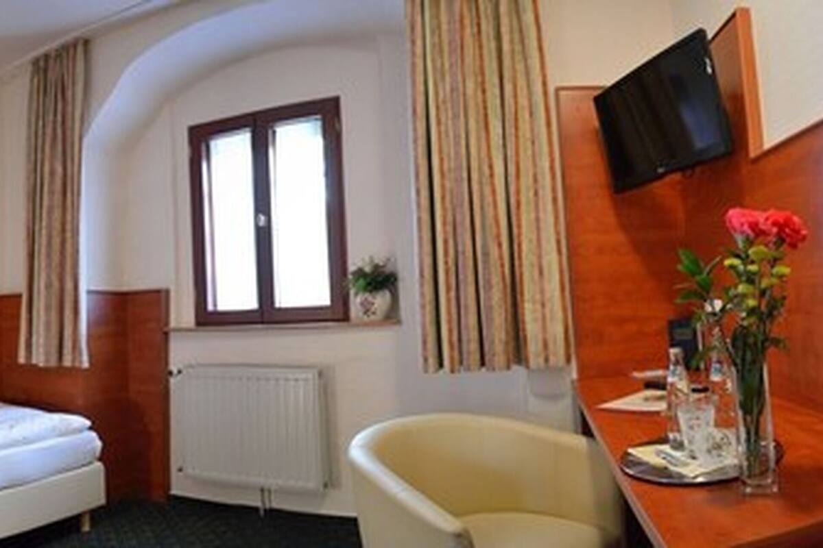 Hotellihuone, jossa on yksi sänky, pieni työpöytä tuolilla, ikkuna verhoilla, patteri ja seinälle asennettu televisio.