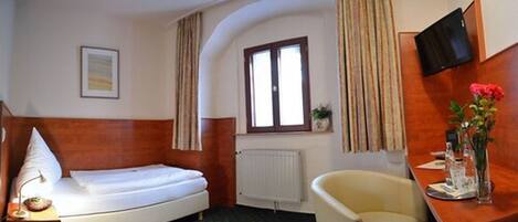 Habitación de hotel con una cama individual, un pequeño escritorio con una silla, una ventana con cortinas, un radiador y un televisor montado en la pared.
