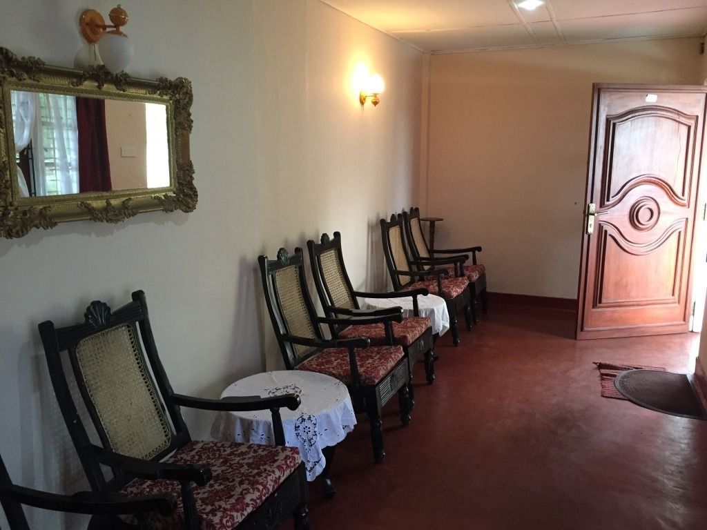 A room with a row of wooden chairs with cushioned seats and backs, a round table with a white tablecloth, a wall-mounted mirror, and a door with a decorative design.