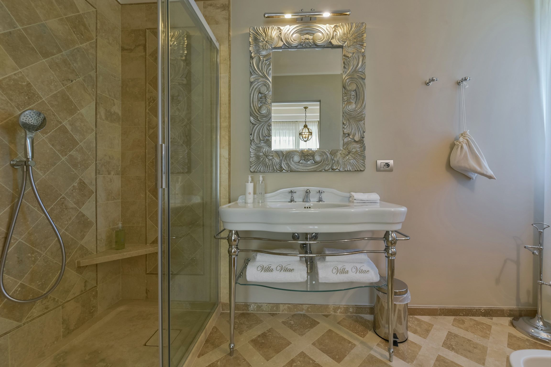 A bathroom with a glass-enclosed shower, a white sink with a glass shelf, and a decorative mirror.
