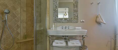 A bathroom with a glass-enclosed shower, a white sink with a glass shelf, and a decorative mirror.