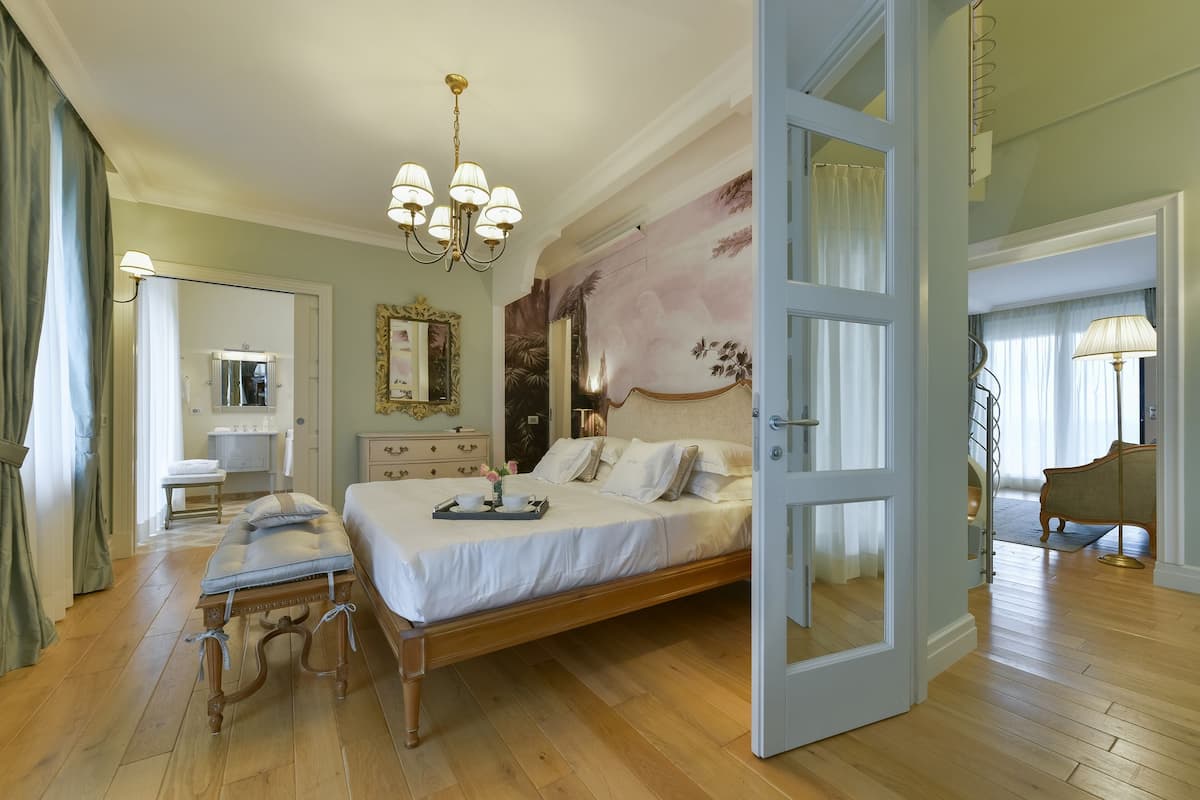 A spacious bedroom with a large bed, a wooden bench, a chandelier, and a view of a staircase.
