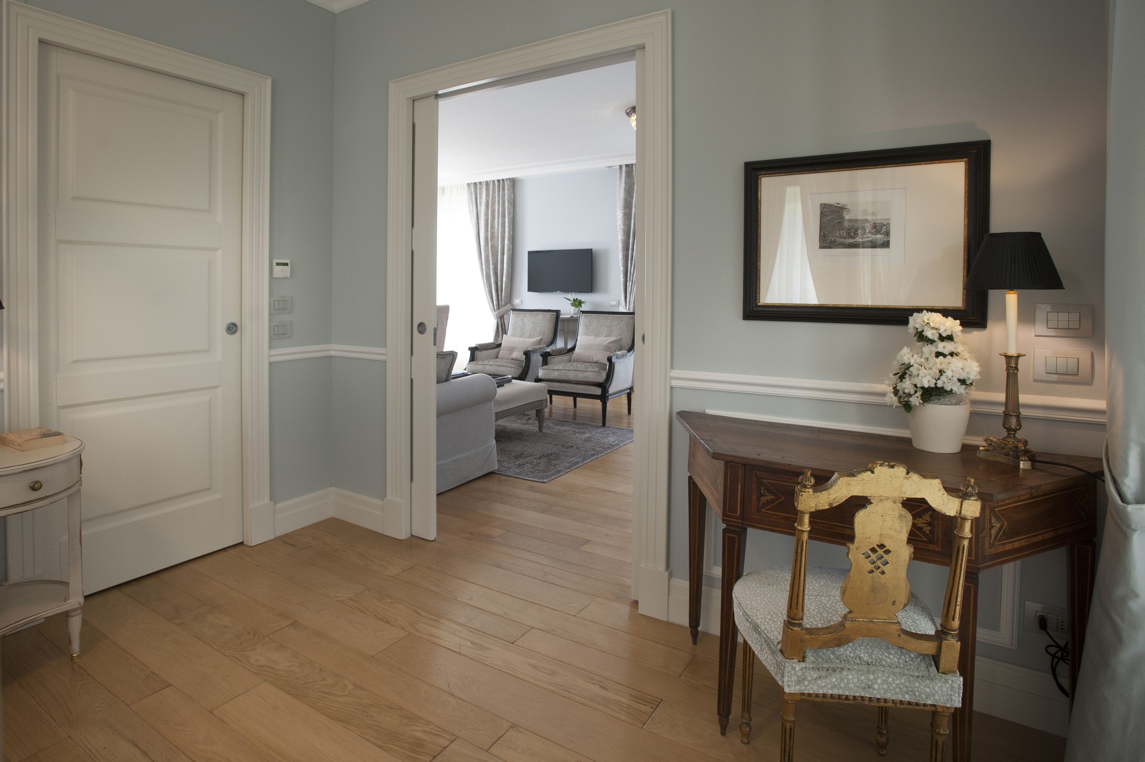 A hallway with a wooden floor, a white door, a small table with a lamp, a framed picture on the wall, and a chair with a cushion.