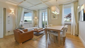 Una sala de estar moderna con un sofá, una mesa de centro, una mesa de comedor y sillas. Hay ventanas amplias con cortinas, un televisor de pantalla plana y una lámpara.