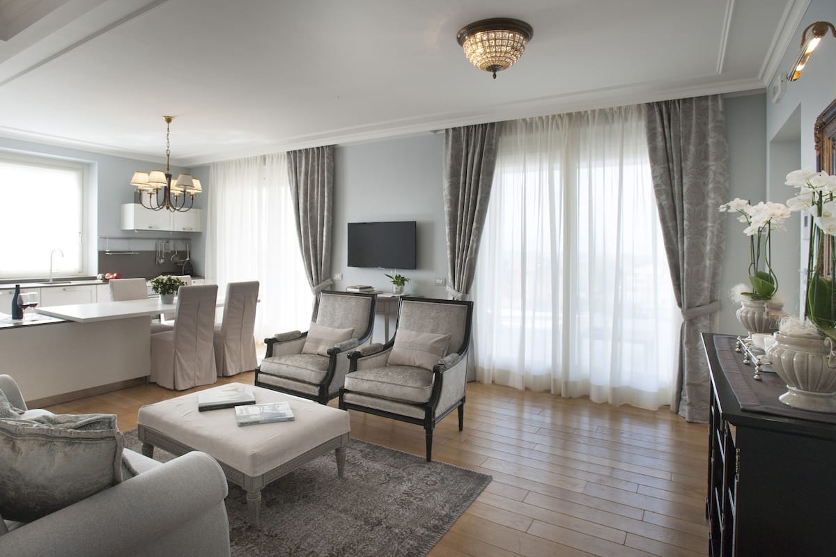 A spacious living room with a sofa, armchairs, a coffee table, and a TV. There is a kitchen area with a dining table and chairs visible through an open door.