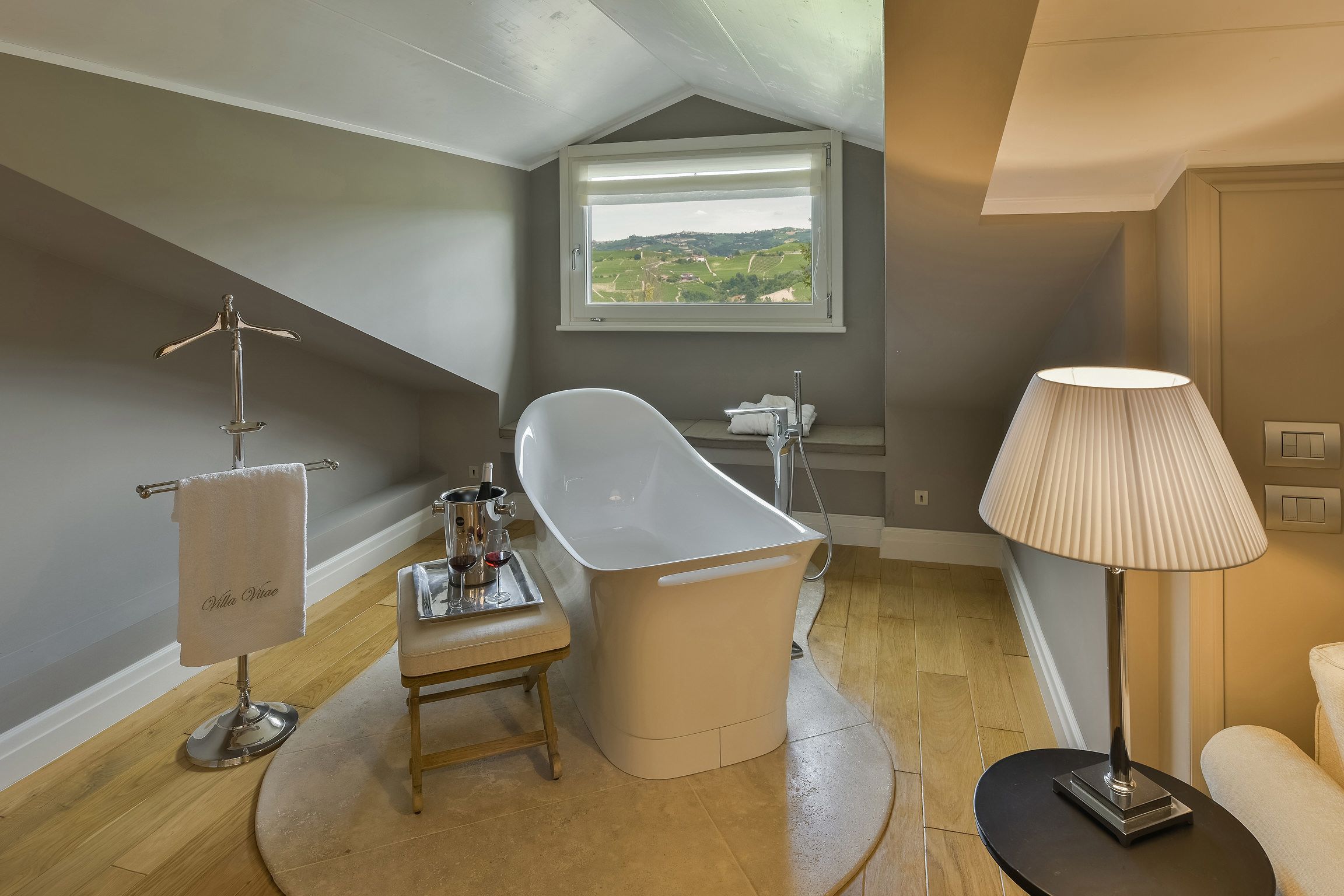 Ein modernes Badezimmer mit einer freistehenden Badewanne, einem Handtuchhalter mit Handtuch, einem kleinen Tisch mit einem Weinglas und einem Fenster mit Blick auf die Landschaft.