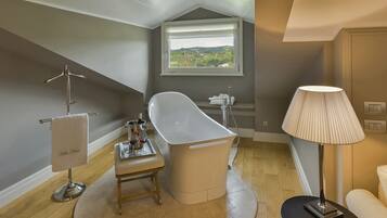 Ein modernes Badezimmer mit einer freistehenden Badewanne, einem Handtuchhalter mit Handtuch, einem kleinen Tisch mit einem Weinglas und einem Fenster mit Blick auf die Landschaft.
