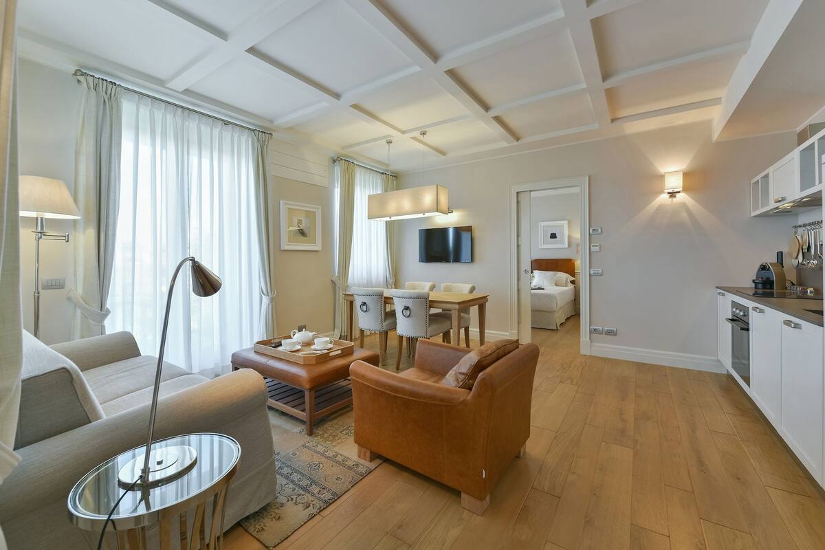 A modern living room with a sofa, a coffee table, a dining area with a table and chairs, a kitchen with cabinets and a stove, and a TV mounted on the wall.