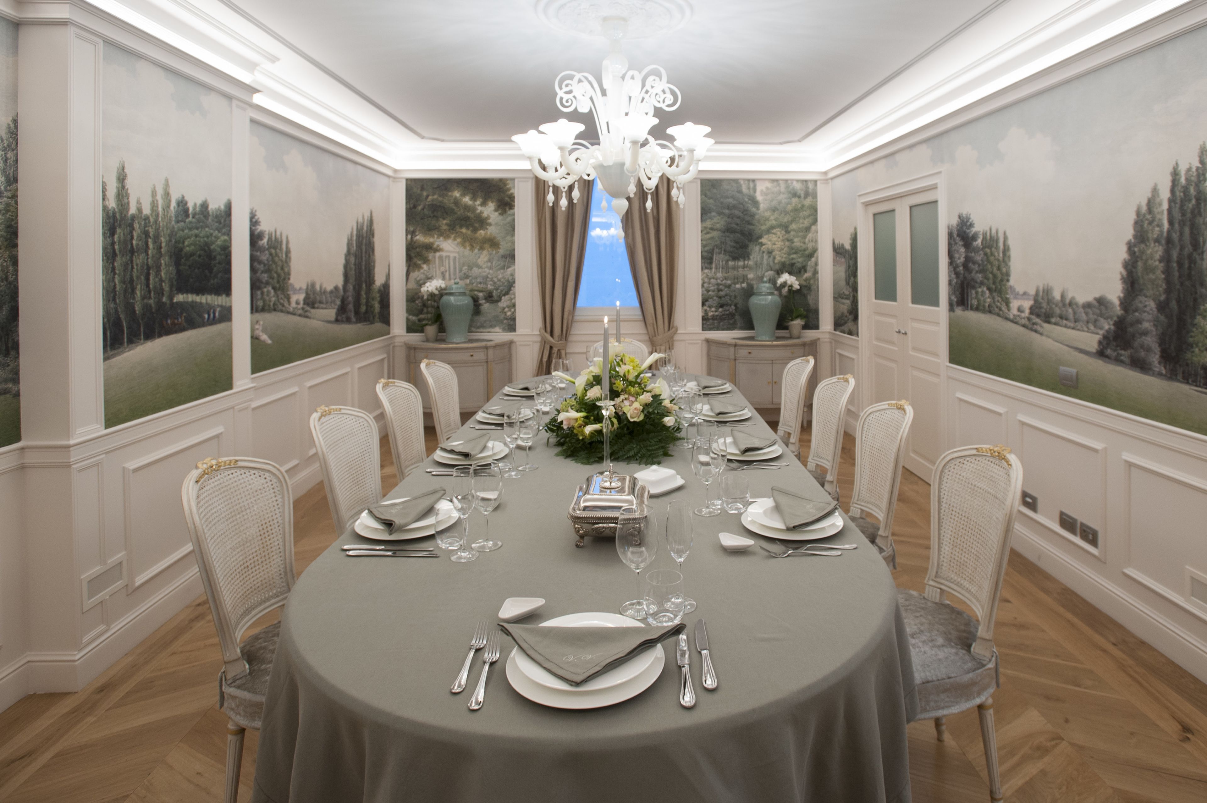 A formal dining room with a long table set for a meal, flanked by two large windows with scenic views.