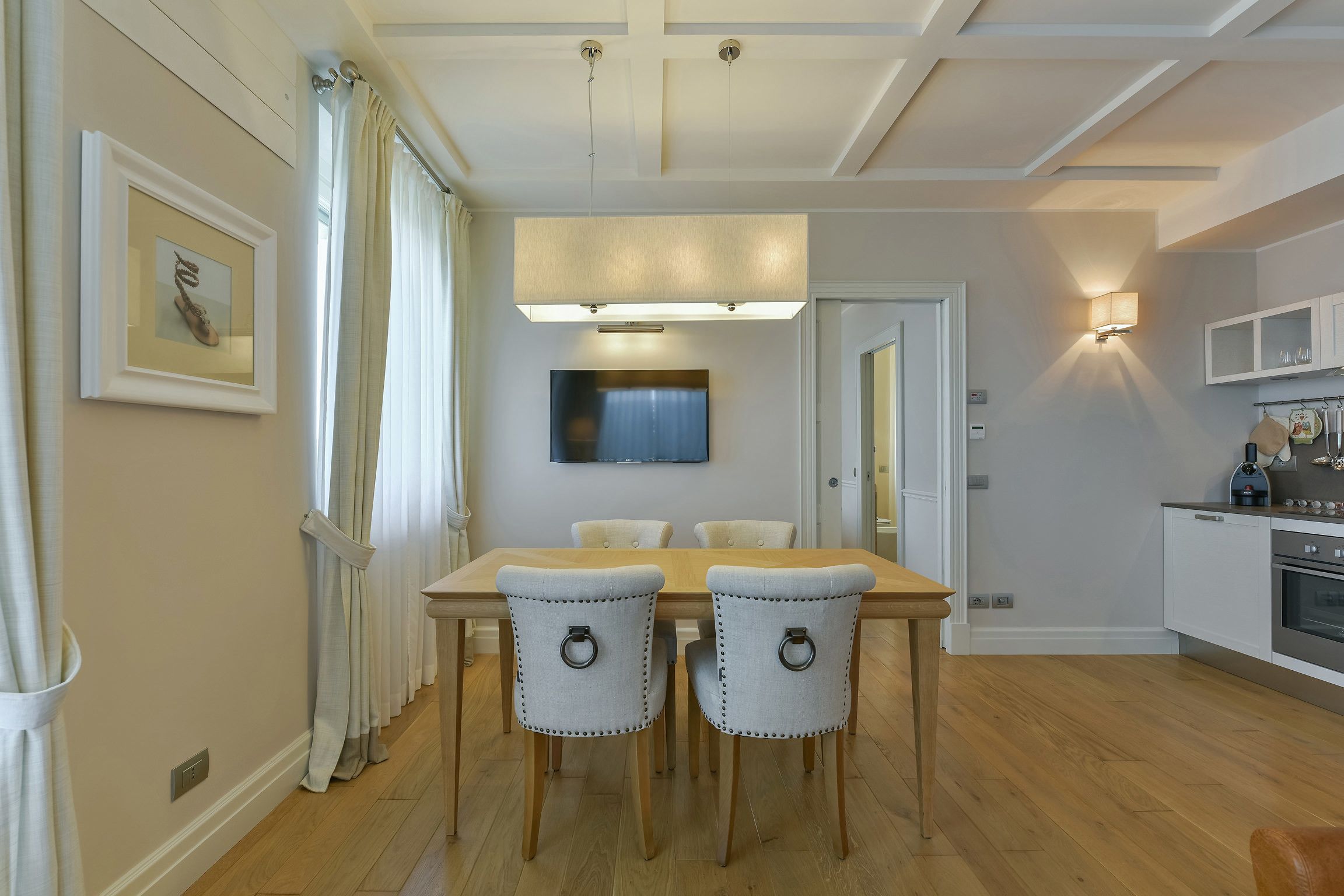 A dining area with a wooden table and four chairs, a flat-screen TV above the table, and a kitchen with modern appliances in the background.