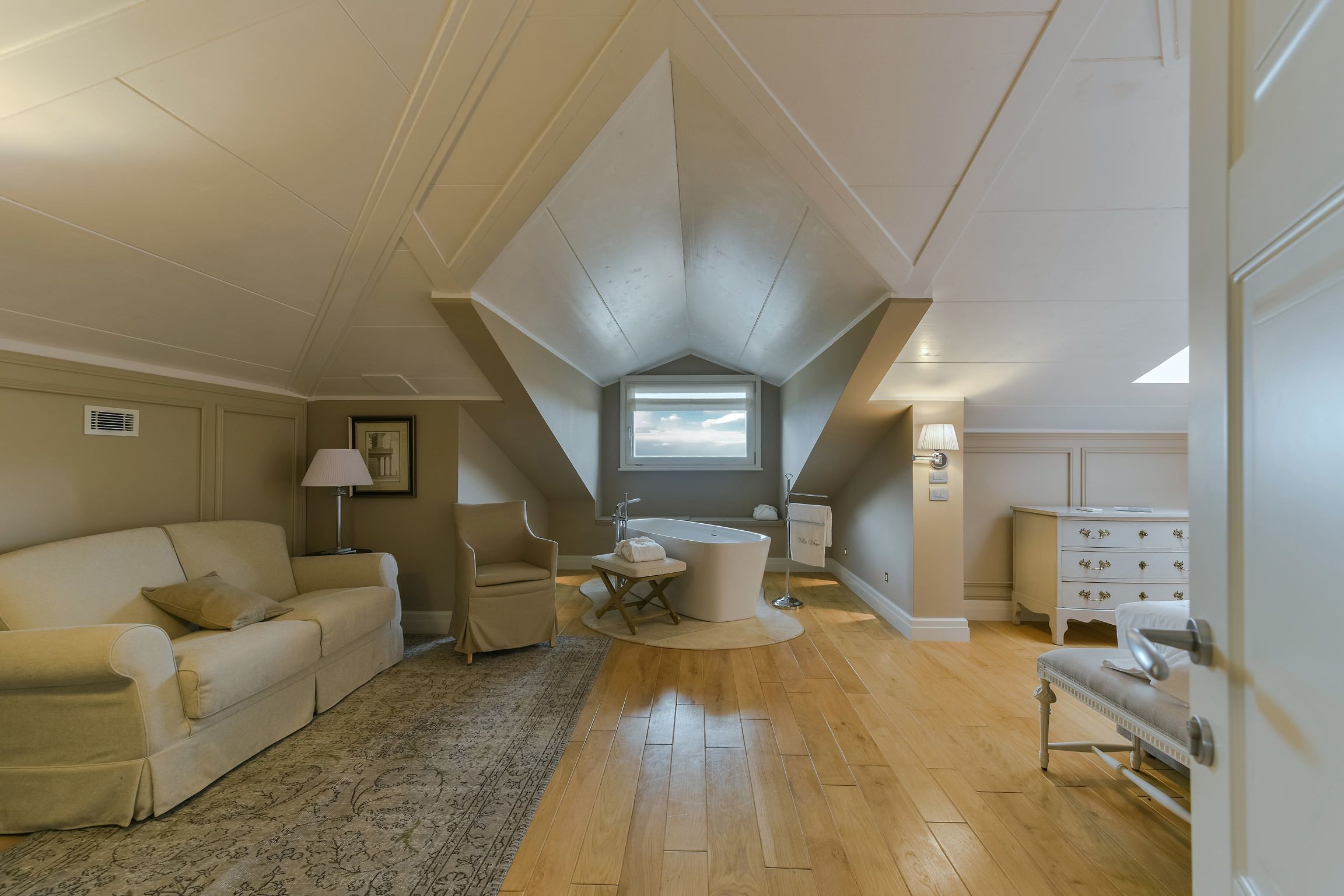 Ruime slaapkamer op zolder met een groot raam, een wit bad, een bank, een stoel en een dressoir.
