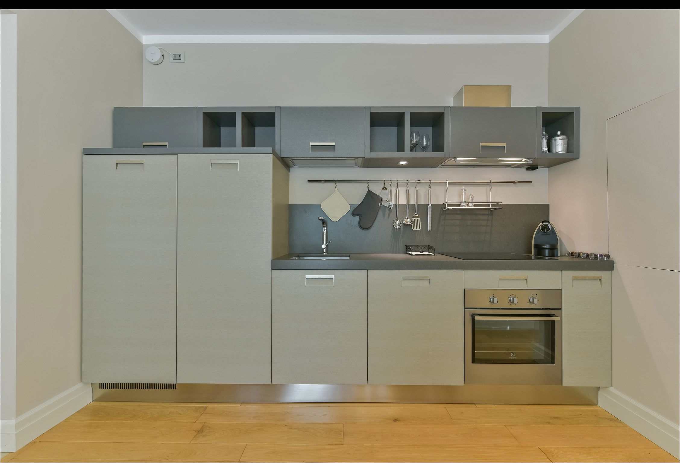 Une cuisine moderne avec des meubles gris, un four encastré et un plan de travail sur lequel se trouvent une bouilloire et une machine à café.