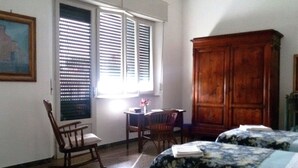 A room with two beds, a wooden wardrobe, a chair, a small table, and a window with blinds. - La Coccinella Guest House (Pisa)