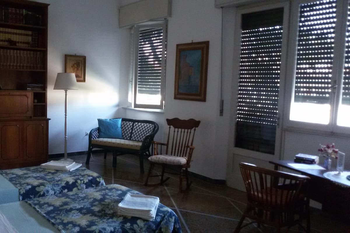 A room with a bed, a chair, a desk, a bookshelf, and two windows with blinds.