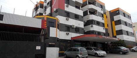 Um hotel com vários andares, fachada vermelha e branca, diversas varandas e estacionamento com carros.