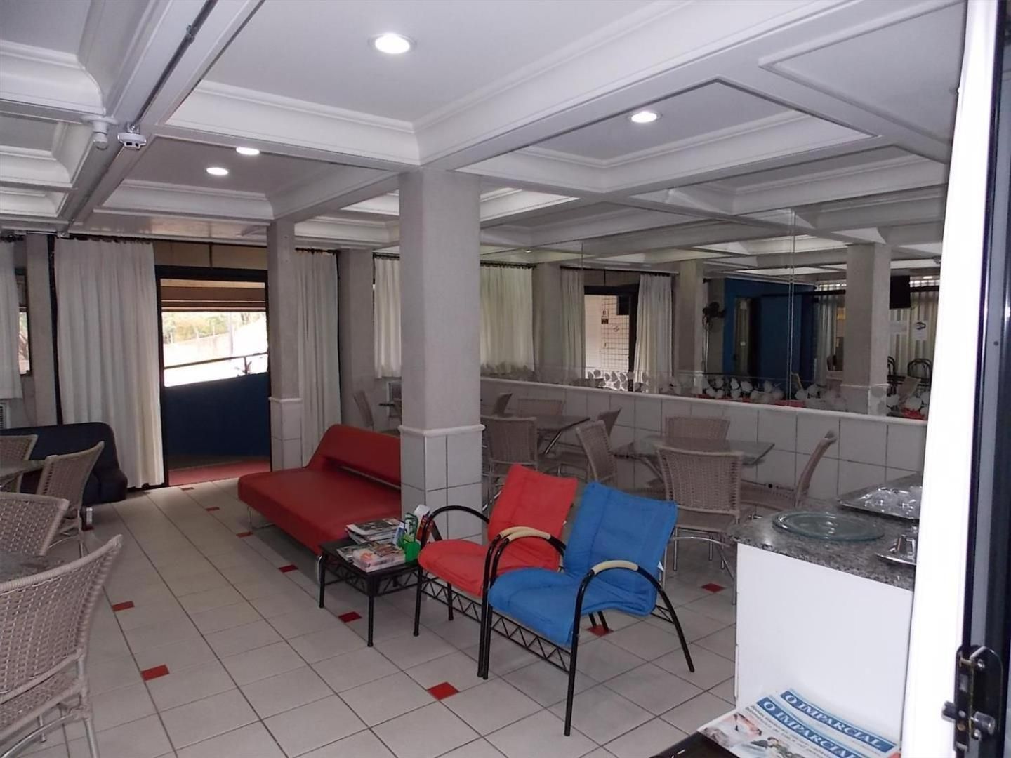 Quarto de hotel com sofá vermelho, poltrona azul e cadeiras de vime. Há uma mesa pequena com revistas e um copo de água. O quarto possui um espelho grande e vista para o exterior através de uma janela com cortinas brancas.