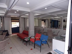 Une chambre d’hôtel avec un canapé rouge, un fauteuil bleu et des chaises en osier. On y trouve une petite table avec des magazines et un verre d’eau. La chambre est dotée d’un grand miroir et offre une vue sur l’extérieur grâce à une fenêtre ornée de rideaux blancs.