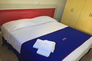 Un lit bien fait, avec du linge de lit blanc rehaussé d’une touche de bleu, une serviette blanche pliée et une armoire jaune aux poignées argentées.