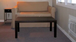 A room with a beige sofa, a small table, a wall-mounted lamp, a framed picture, and a window with blinds. - Balm Beach Resort & Motel (Tiny)