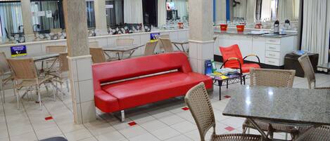 Un vestíbulo de hotel con un sofá rojo, sillas de mimbre, una mesa de granito y una zona de cocina con un fregadero y encimera.
