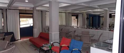 A hotel room with a red sofa, blue armchair, and wicker chairs. There is a dining area with a glass table and chairs. The room has a tiled floor and a large mirror on one wall.